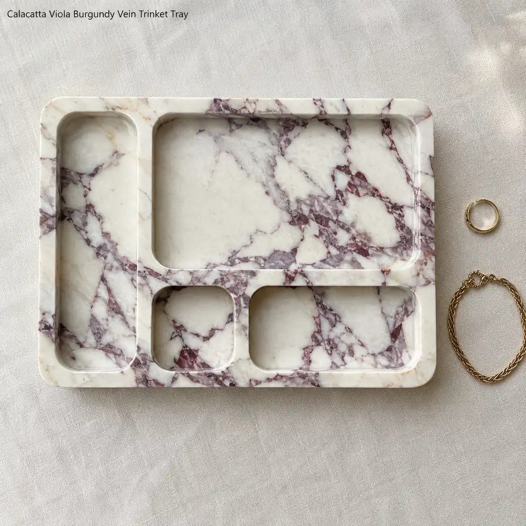 Overhead Calacatta Viola trinket tray on pale linen with carved compartments and burgundy stone movement.