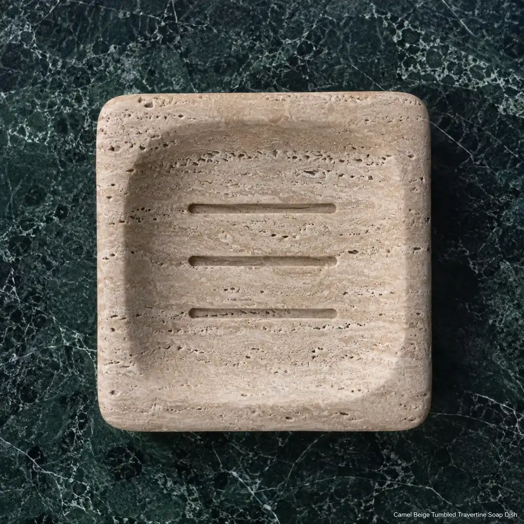Camel beige travertine soap dish on forest-green stone, overhead view showing tumbled rim and drainage grooves.