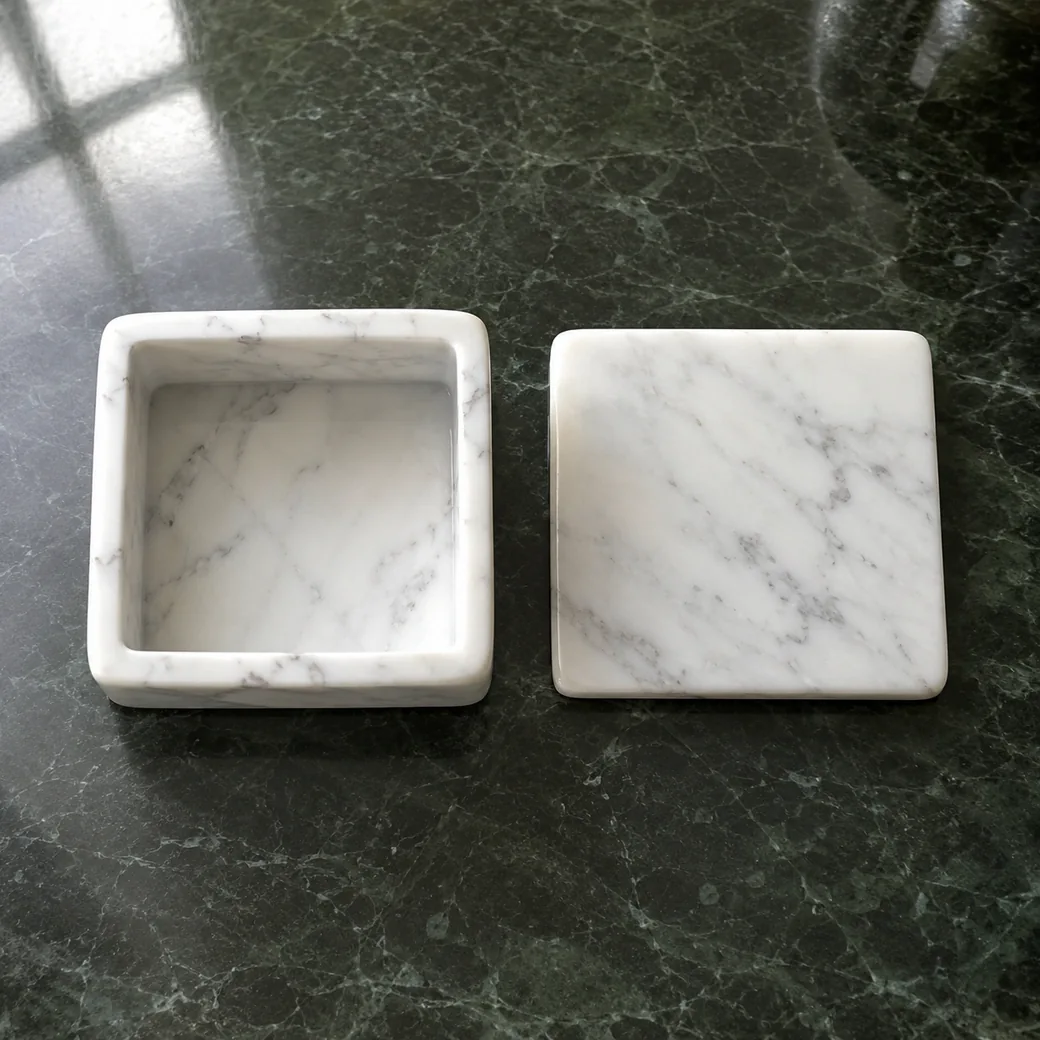 Open Carrara coaster box viewed overhead on dark green stone, with lid beside it and polished square interior visible.