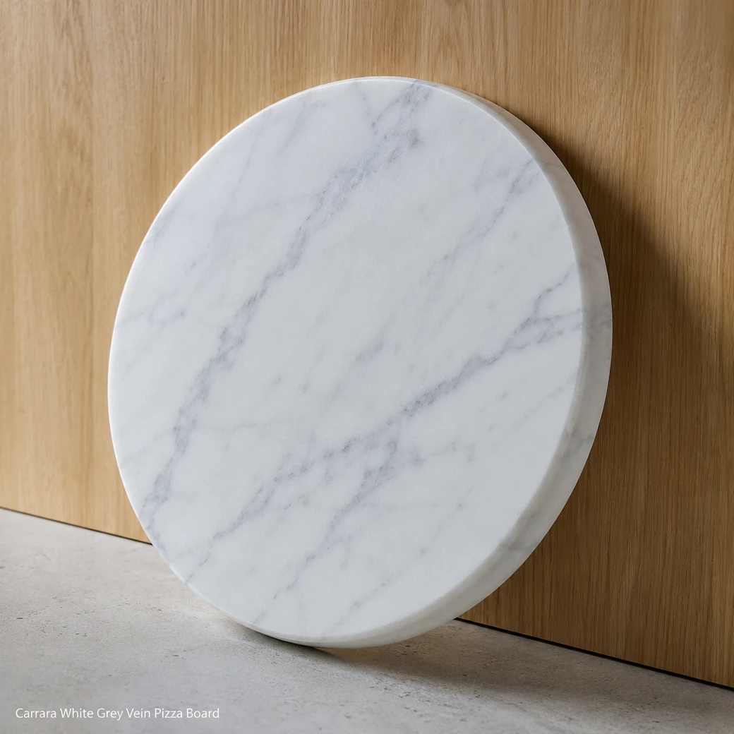 Carrara pizza board leaning against pale oak backsplash, low side view showing honed face, grey veins and 2 cm edge.