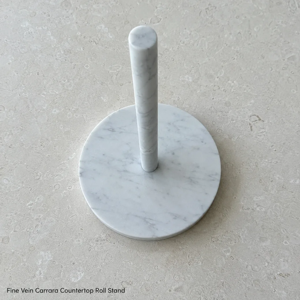 Carrara paper towel holder on pale limestone, overhead diagonal view showing base geometry and centered spindle.