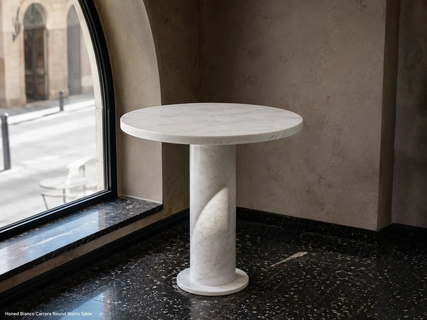 Honed Carrara bistro table beside an arched cafe window, photographed low on dark terrazzo
